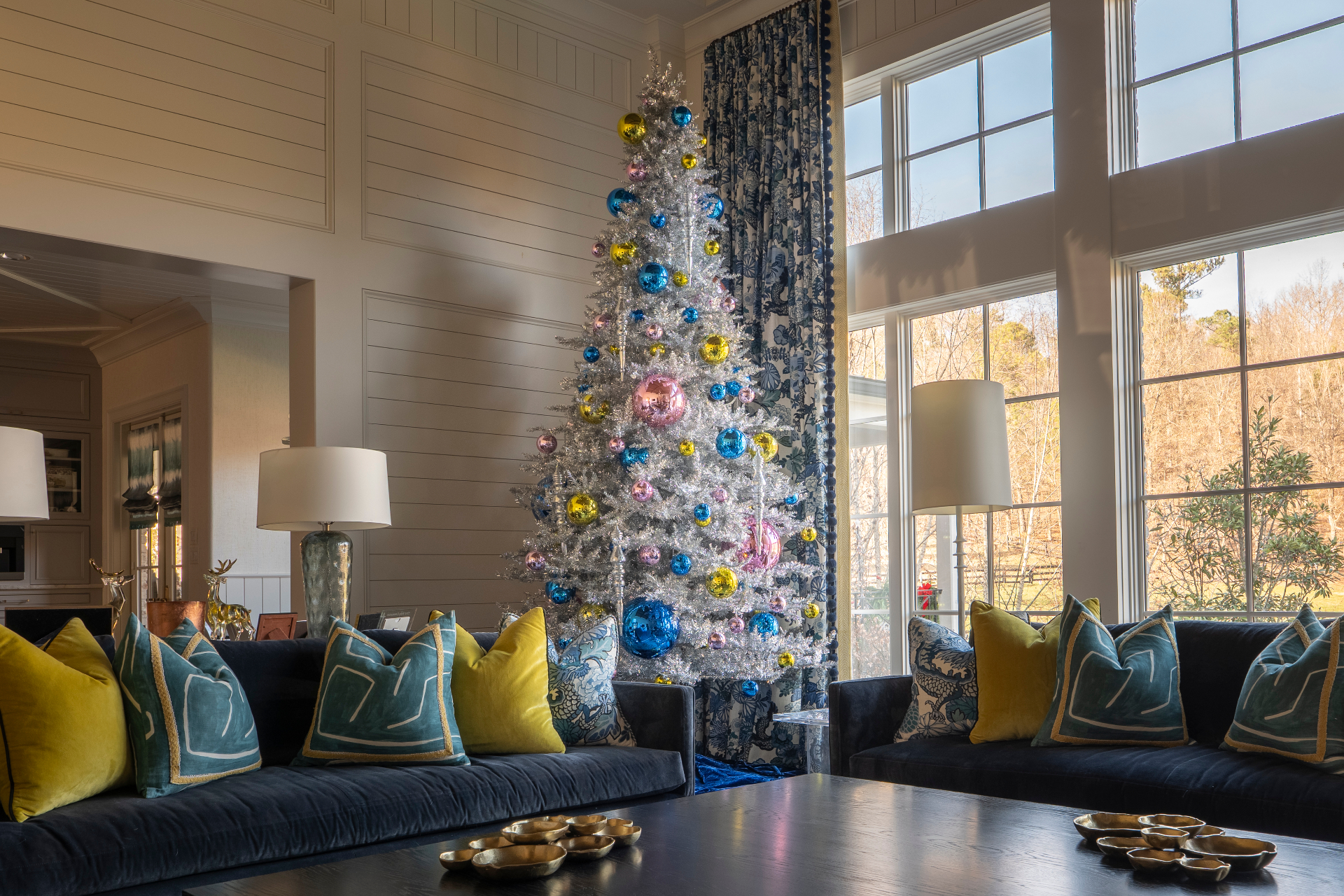 A bright silver tinsel Christmas tree with bold blue, pink, and gold ornaments in a sunlit living room