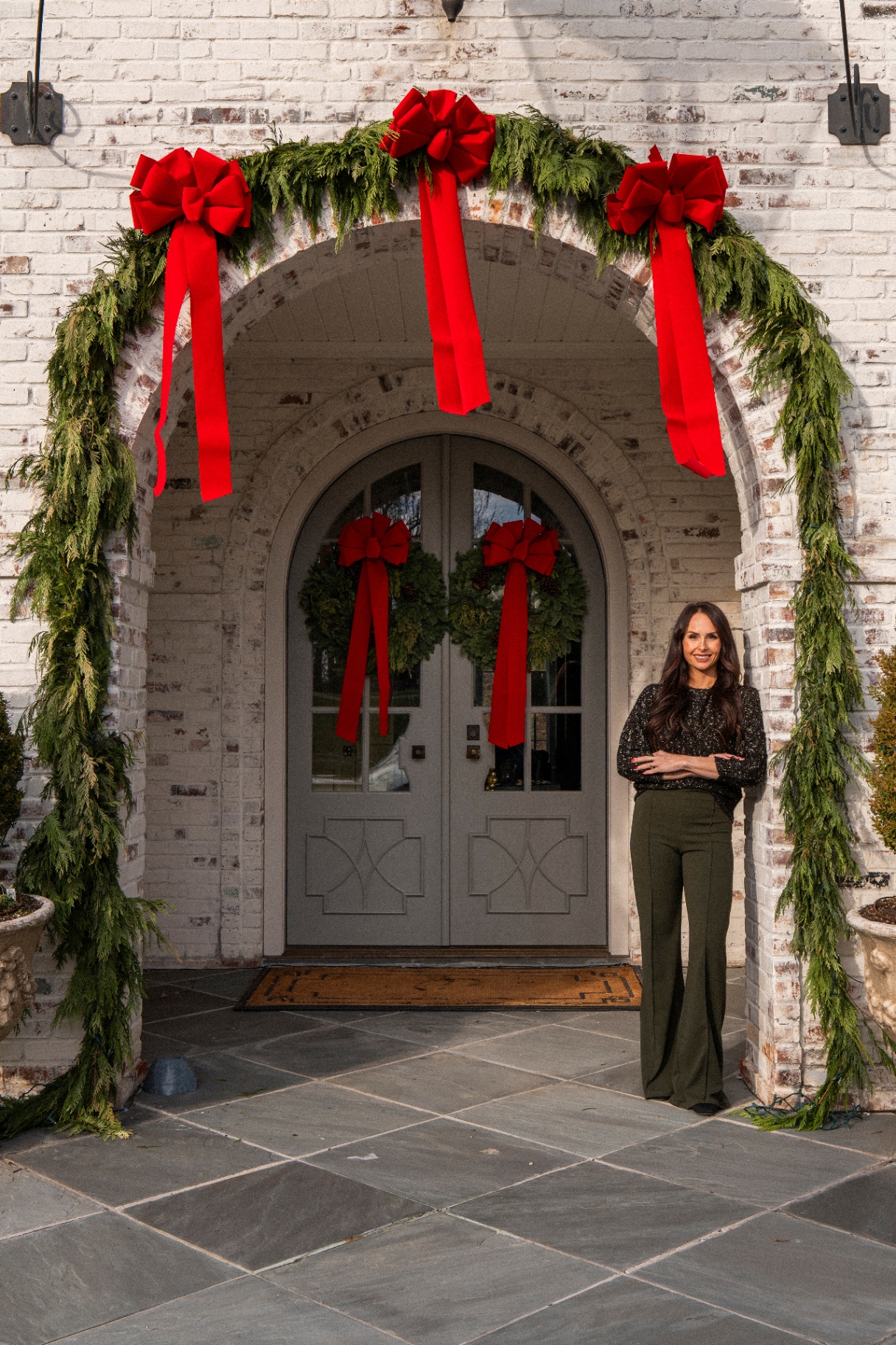Jen standing at an arched front entrance decorated with fresh garland and red velvet bows