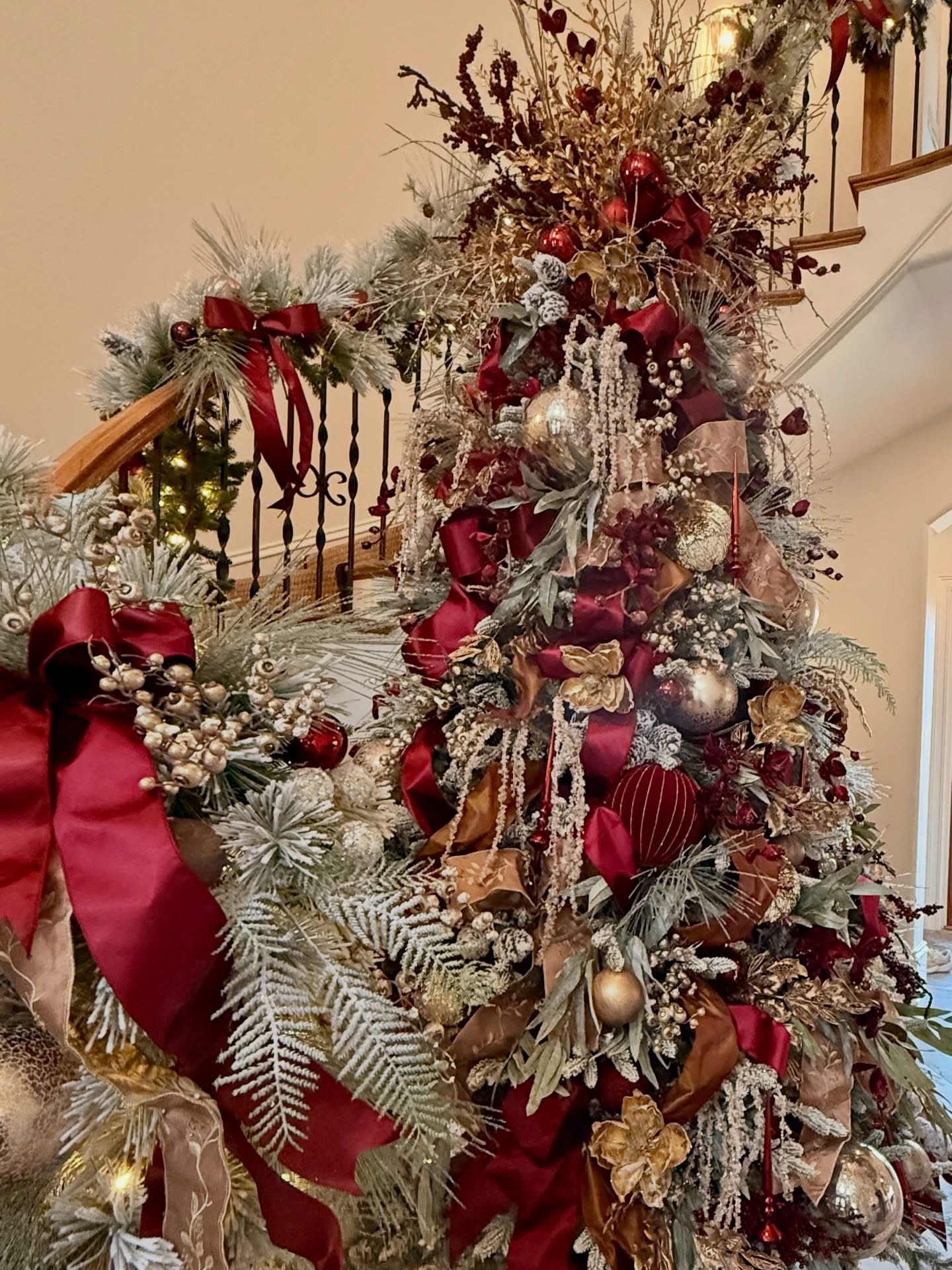 Towering flocked Christmas tree dressed in burgundy ribbon, gold magnolia, and mercury ornaments beside a wrought-iron staircase
