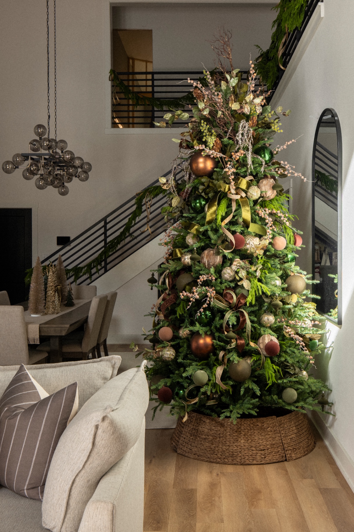 A grand Christmas tree with copper, sage, and blush ornaments, olive and gold ribbon, nestled beside a modern staircase in a luxury home